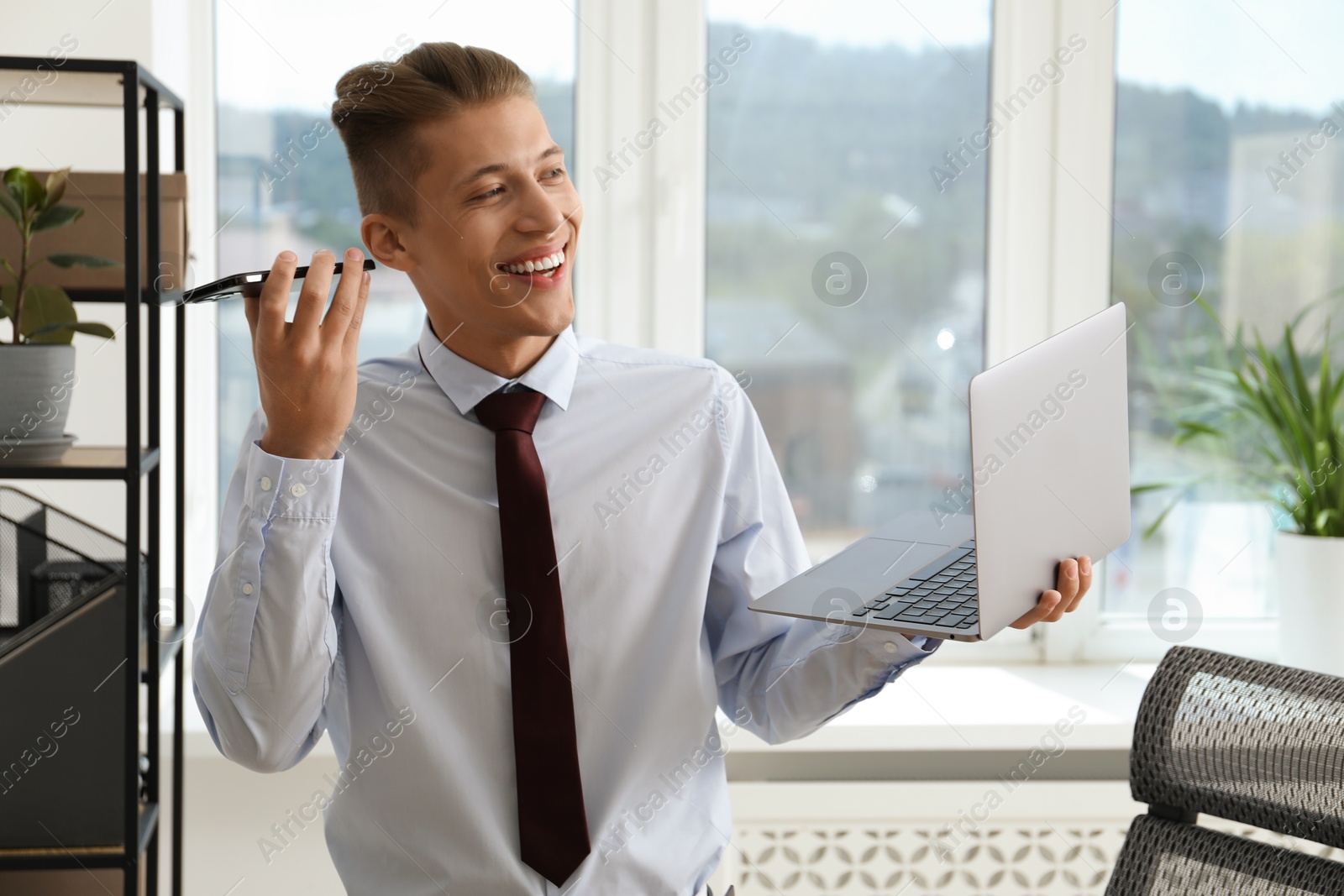 Photo of Young man with laptop listening to voice message via smartphone in office