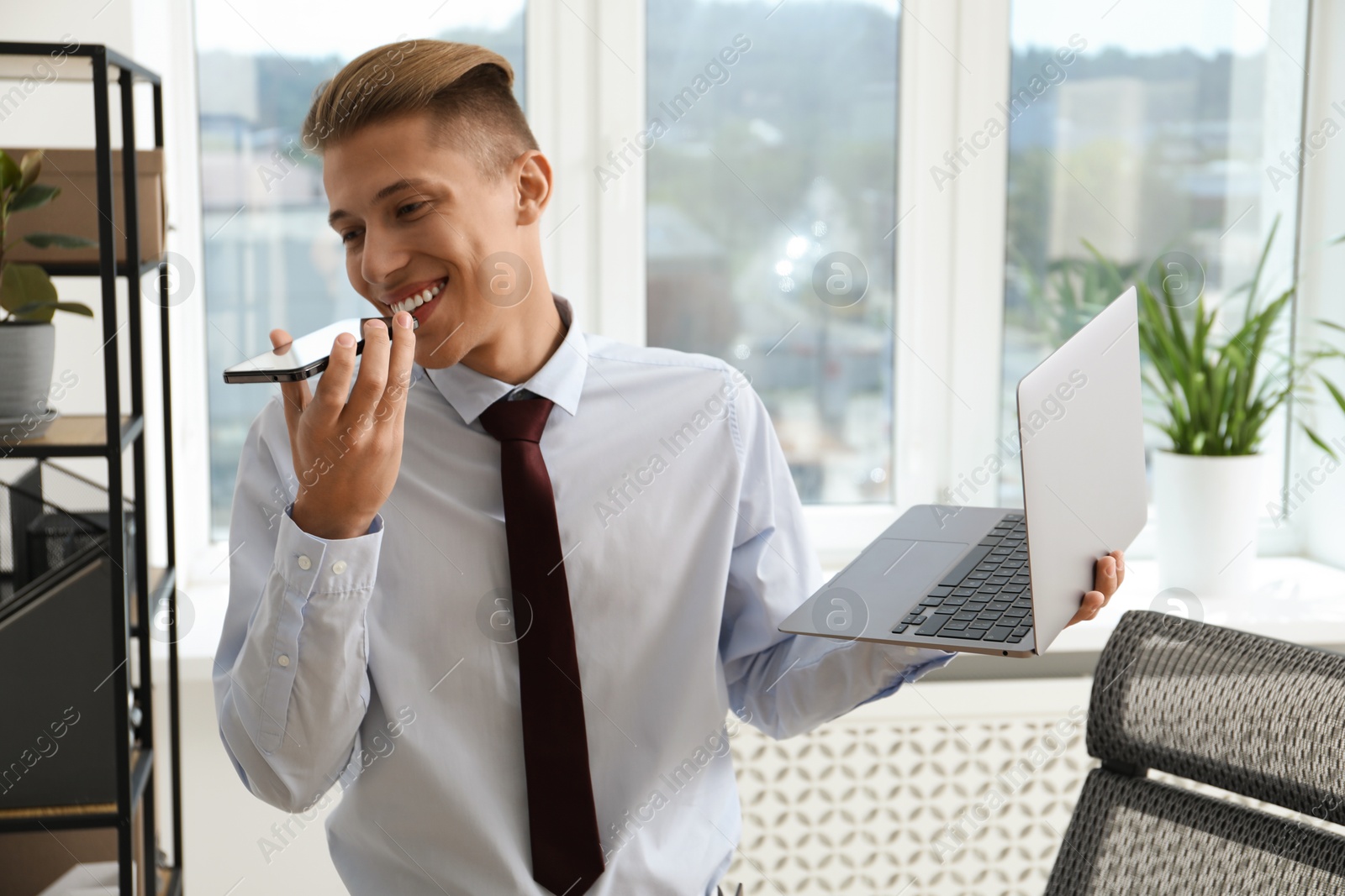 Photo of Young man with laptop listening to voice message via smartphone in office