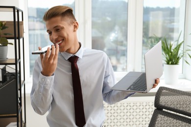 Photo of Young man with laptop listening to voice message via smartphone in office