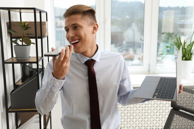 Young man with laptop recording voice message via smartphone in office