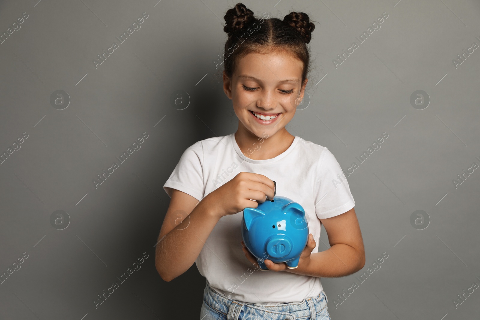 Photo of Pocket money. Cute girl putting coins into piggy bank on grey background