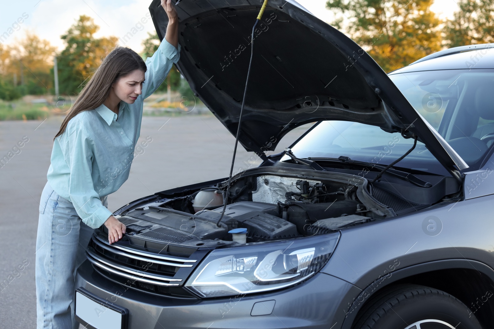 Photo of Stressed woman looking under hood of broken car outdoors