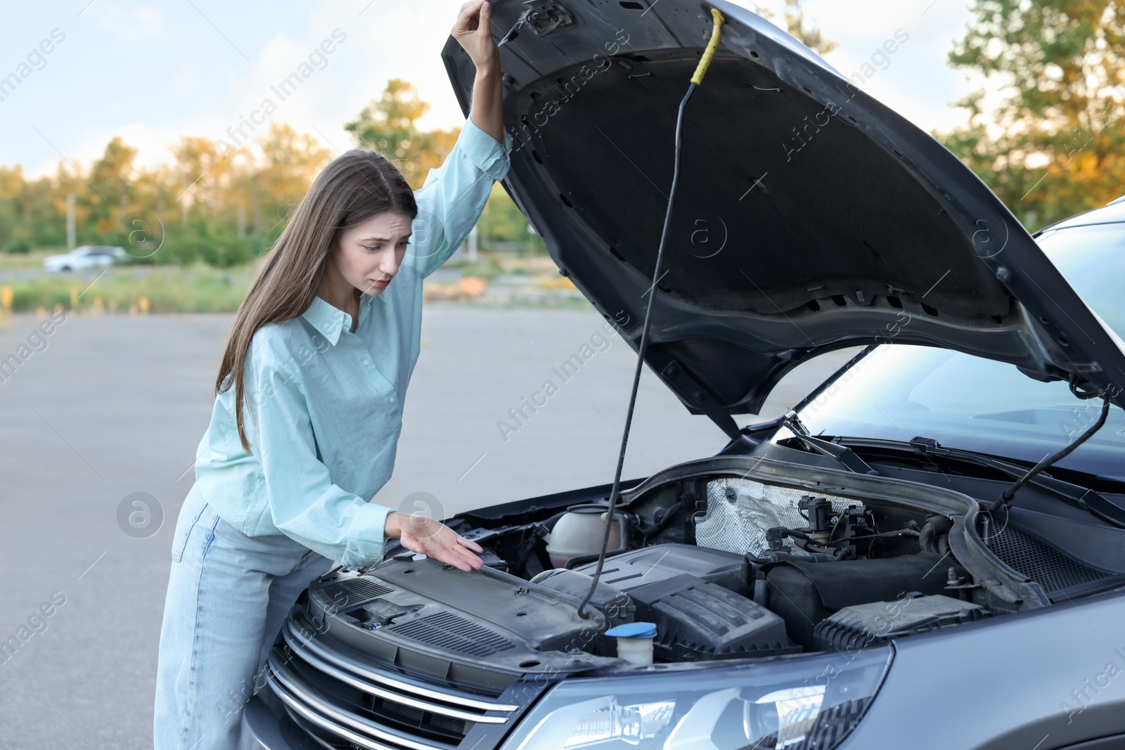 Photo of Stressed woman looking under hood of broken car outdoors