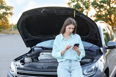 Stressed woman using smartphone near broken car outdoors