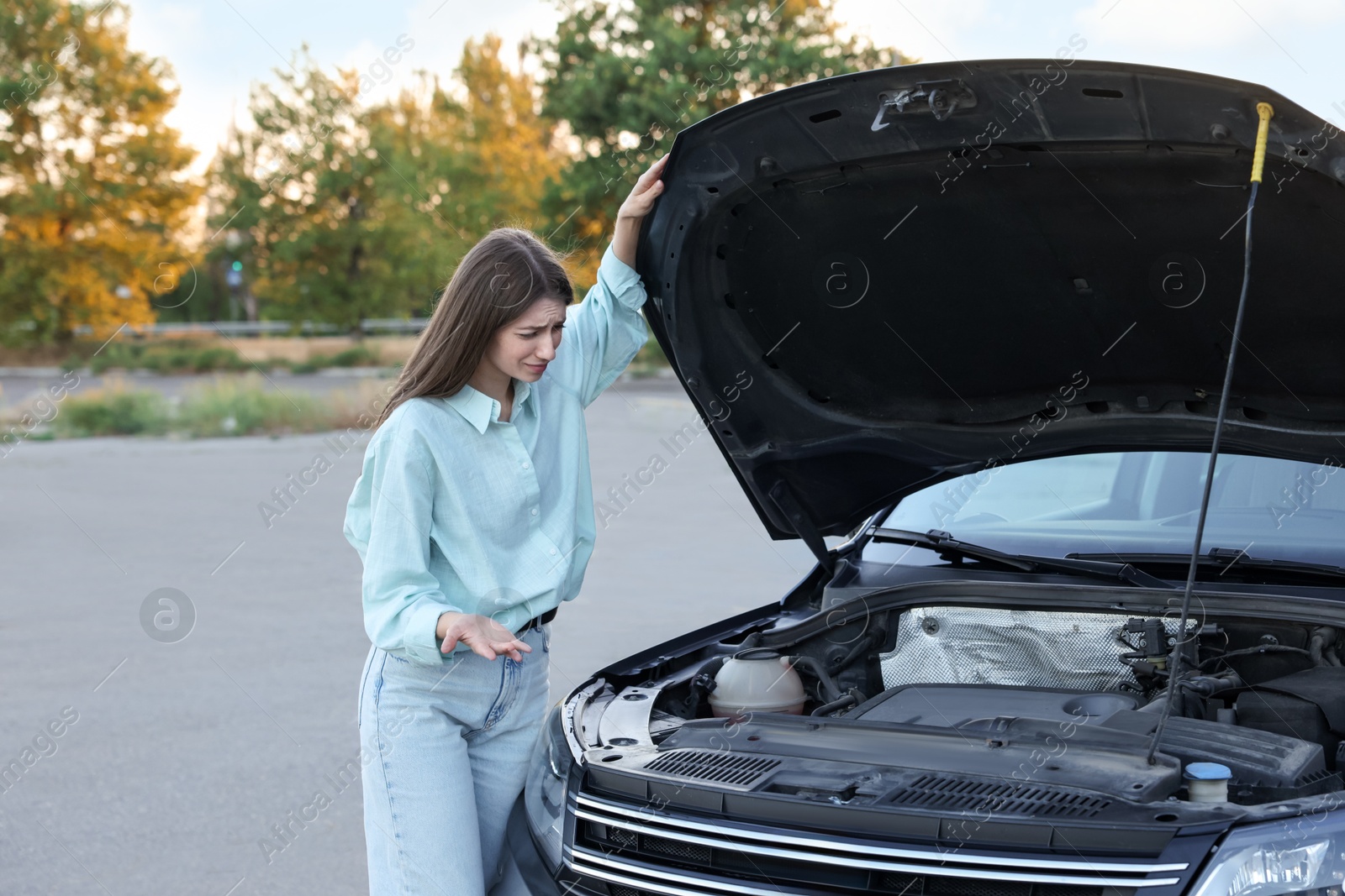 Photo of Stressed woman looking under hood of broken car outdoors