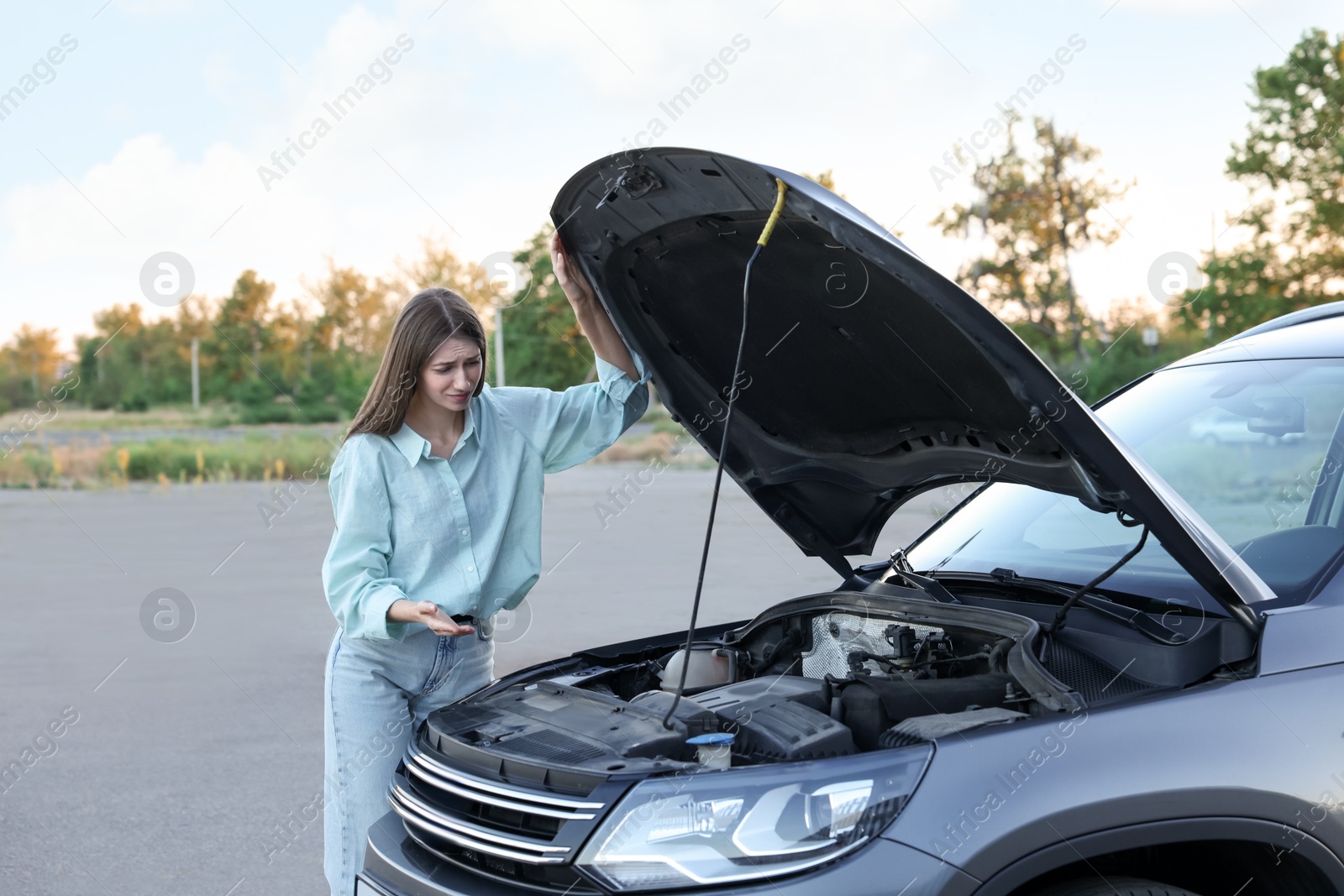 Photo of Stressed woman looking under hood of broken car outdoors