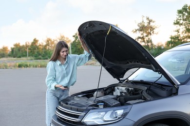 Stressed woman looking under hood of broken car outdoors