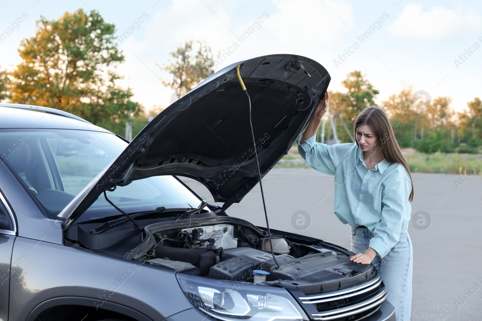 Photo of Stressed woman looking under hood of broken car outdoors