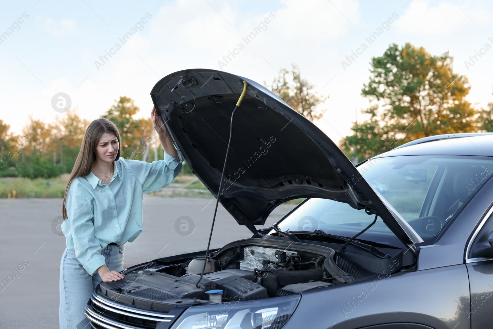 Photo of Stressed woman looking under hood of broken car outdoors