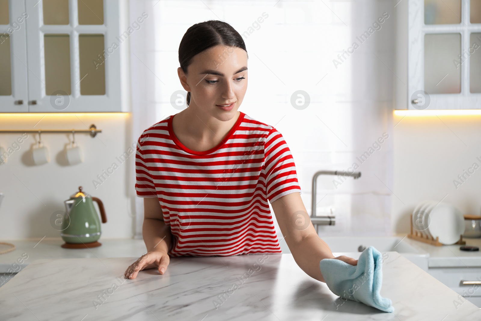 Photo of Beautiful young woman wiping white marble table with rag in kitchen