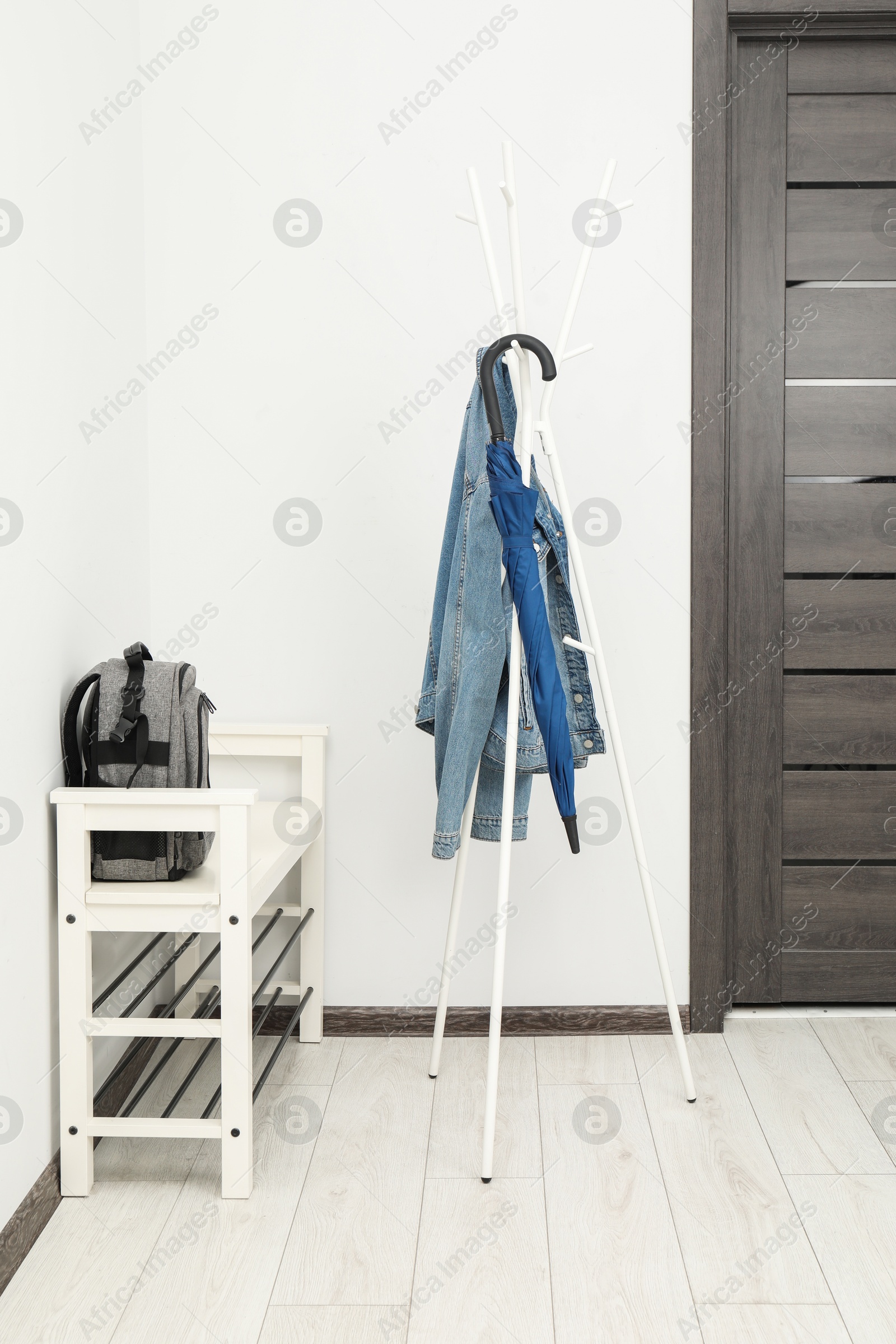 Photo of Blue umbrella and denim jacket on clothing rack near shoe storage bench with backpack in hallway