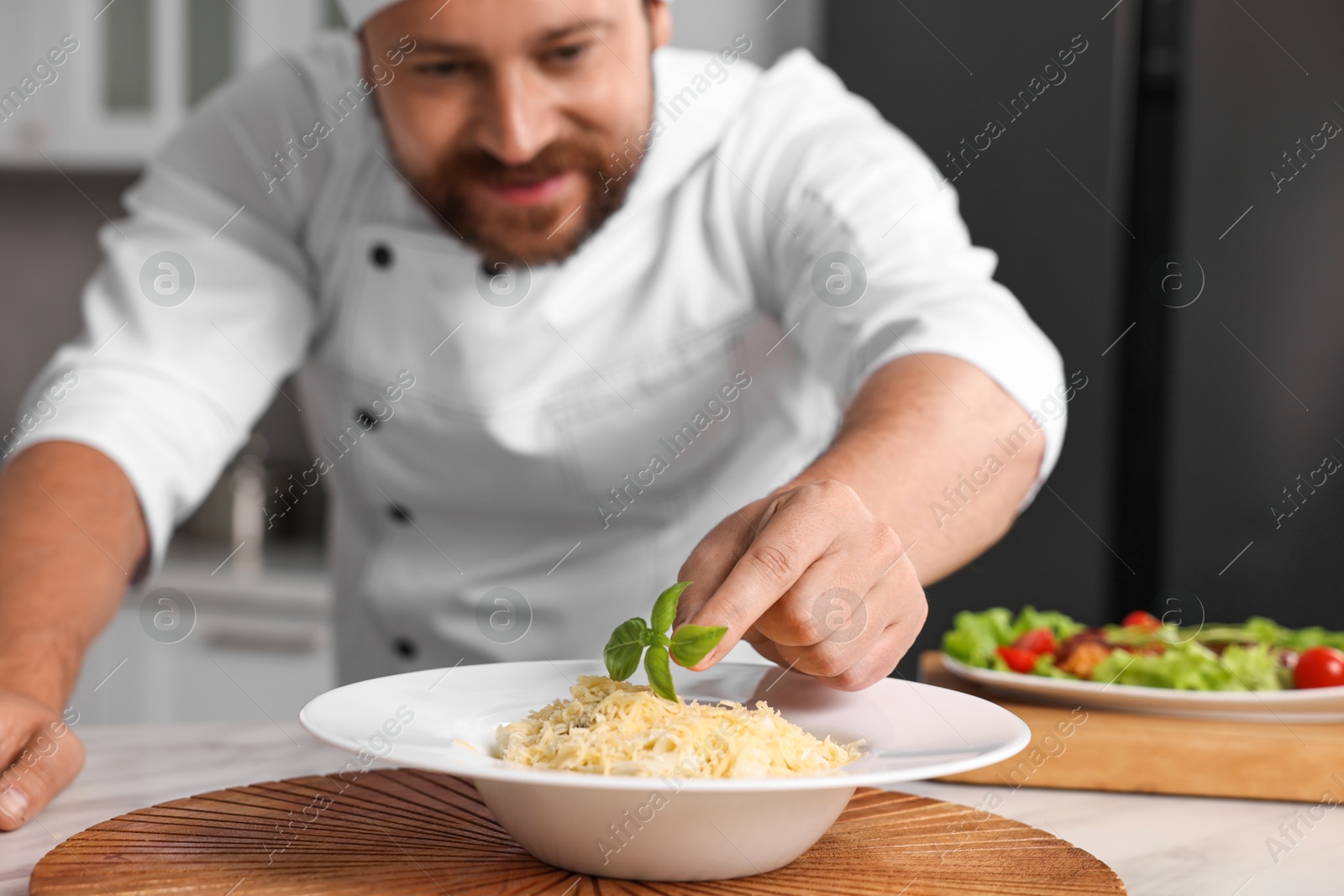 Photo of Professional chef decorating delicious pasta with basil at white marble table in kitchen, selective focus