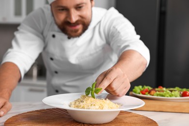 Professional chef decorating delicious pasta with basil at white marble table in kitchen, selective focus