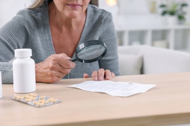 Photo of Senior woman with magnifying glass reading medicine instruction at table indoors, closeup