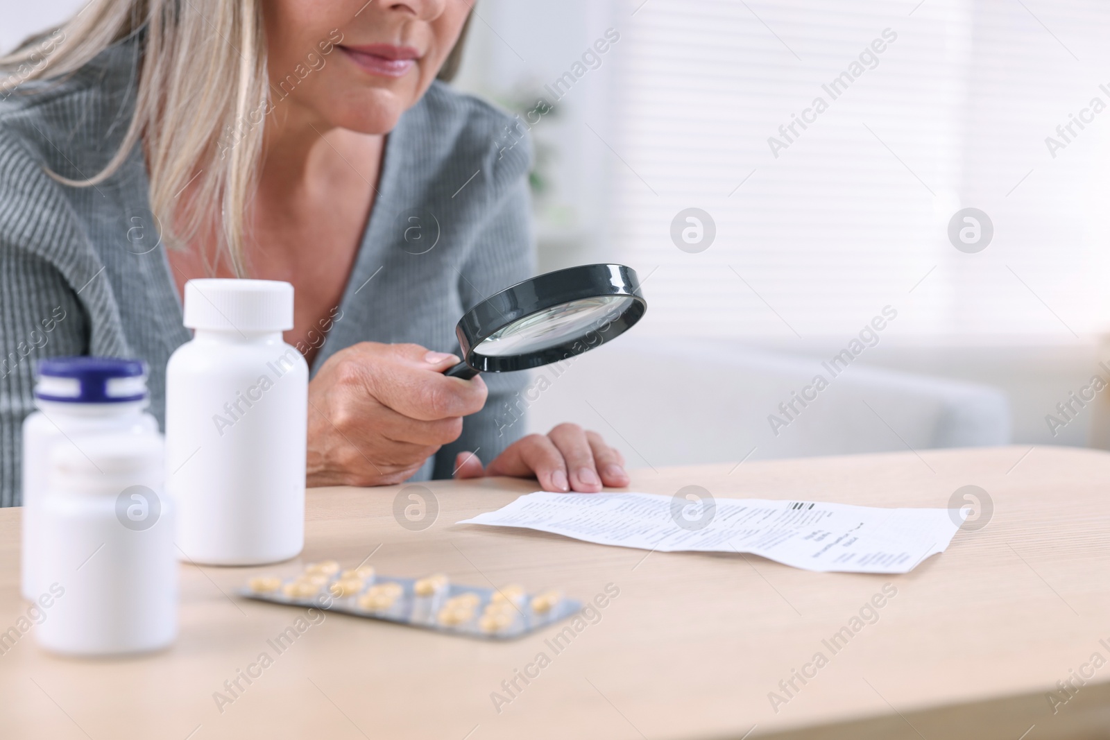 Photo of Senior woman with magnifying glass reading medicine instruction at table indoors, closeup