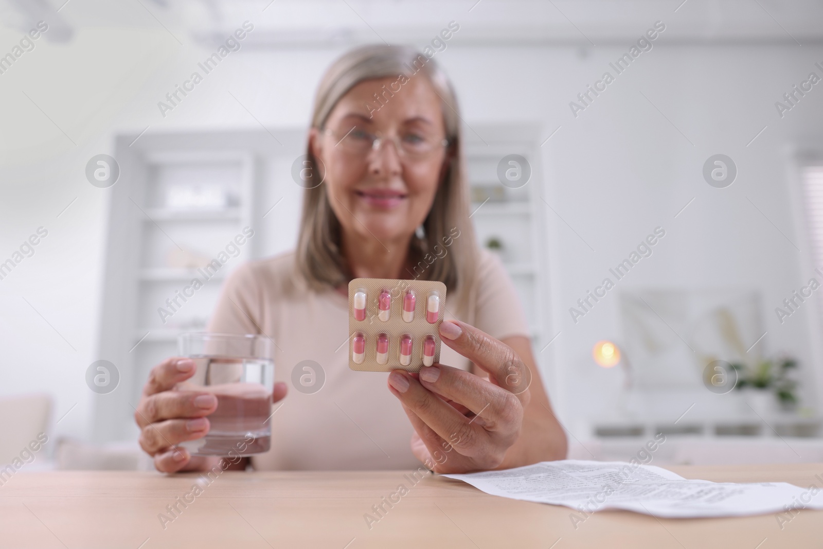 Photo of Senior woman with pills and glass of water at table indoors, selective focus