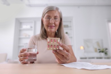 Senior woman with pills and glass of water at table indoors, selective focus