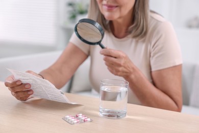 Senior woman with magnifying glass reading medicine instruction at table indoors, closeup