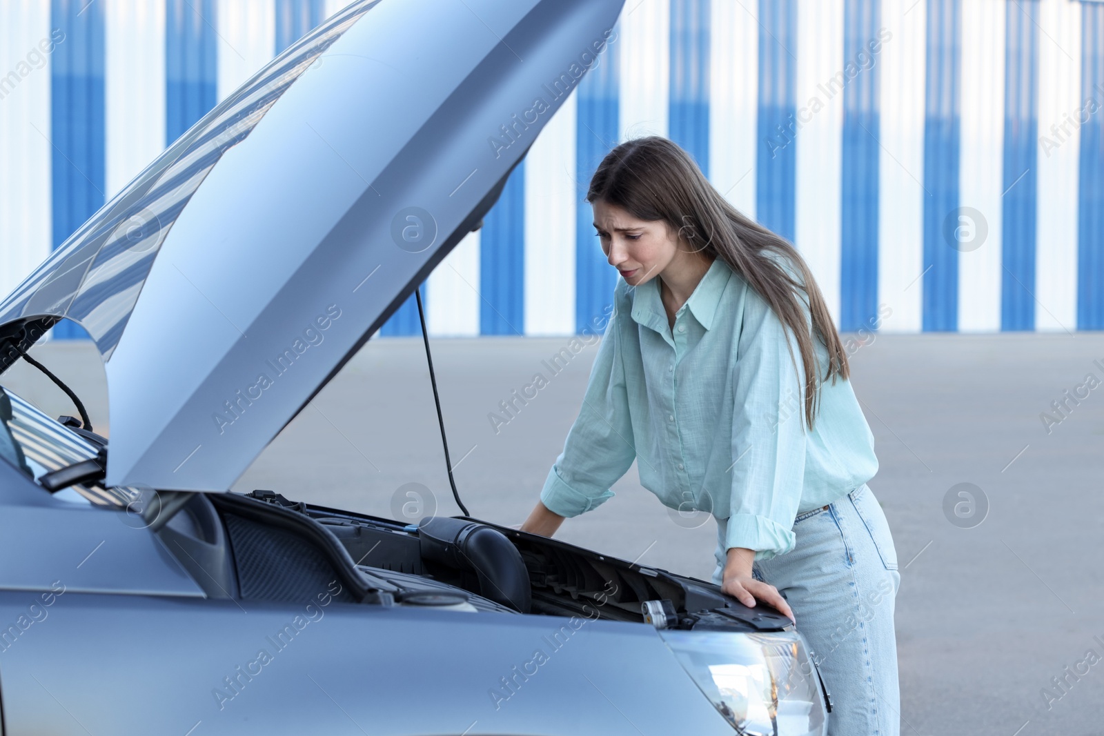 Photo of Stressed woman looking under hood of broken car outdoors