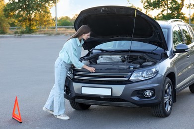 Stressed woman looking under hood of broken car outdoors