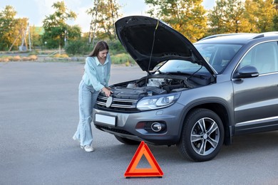 Photo of Stressed woman looking under hood of broken car outdoors