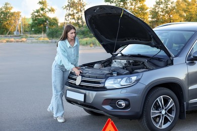 Photo of Stressed woman looking under hood of broken car outdoors