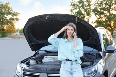 Stressed woman talking on phone near broken car outdoors