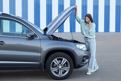 Stressed woman talking on phone near broken car outdoors