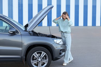 Stressed woman looking under hood of broken car outdoors