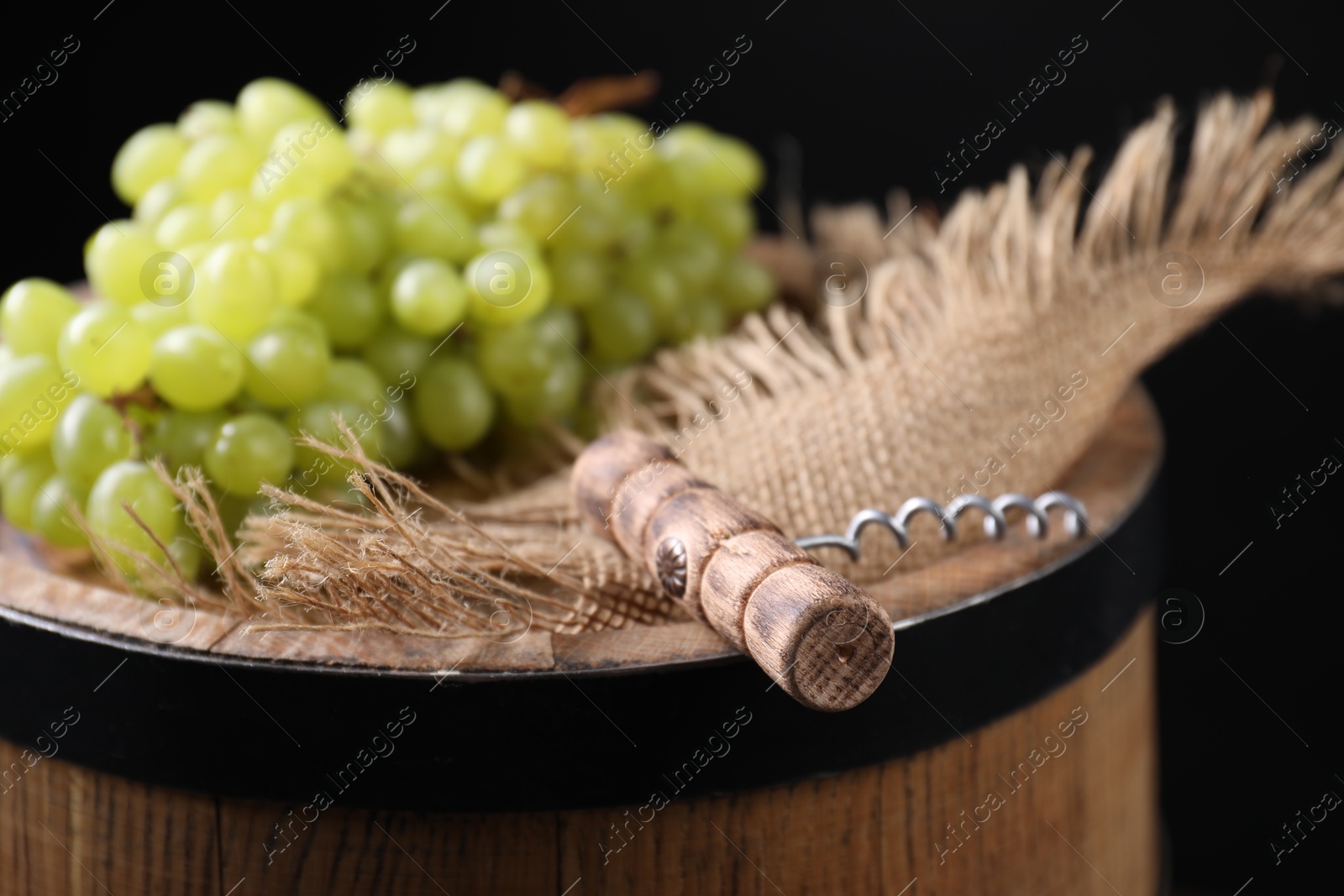 Photo of Corkscrew and grapes on wooden barrel against black background, closeup