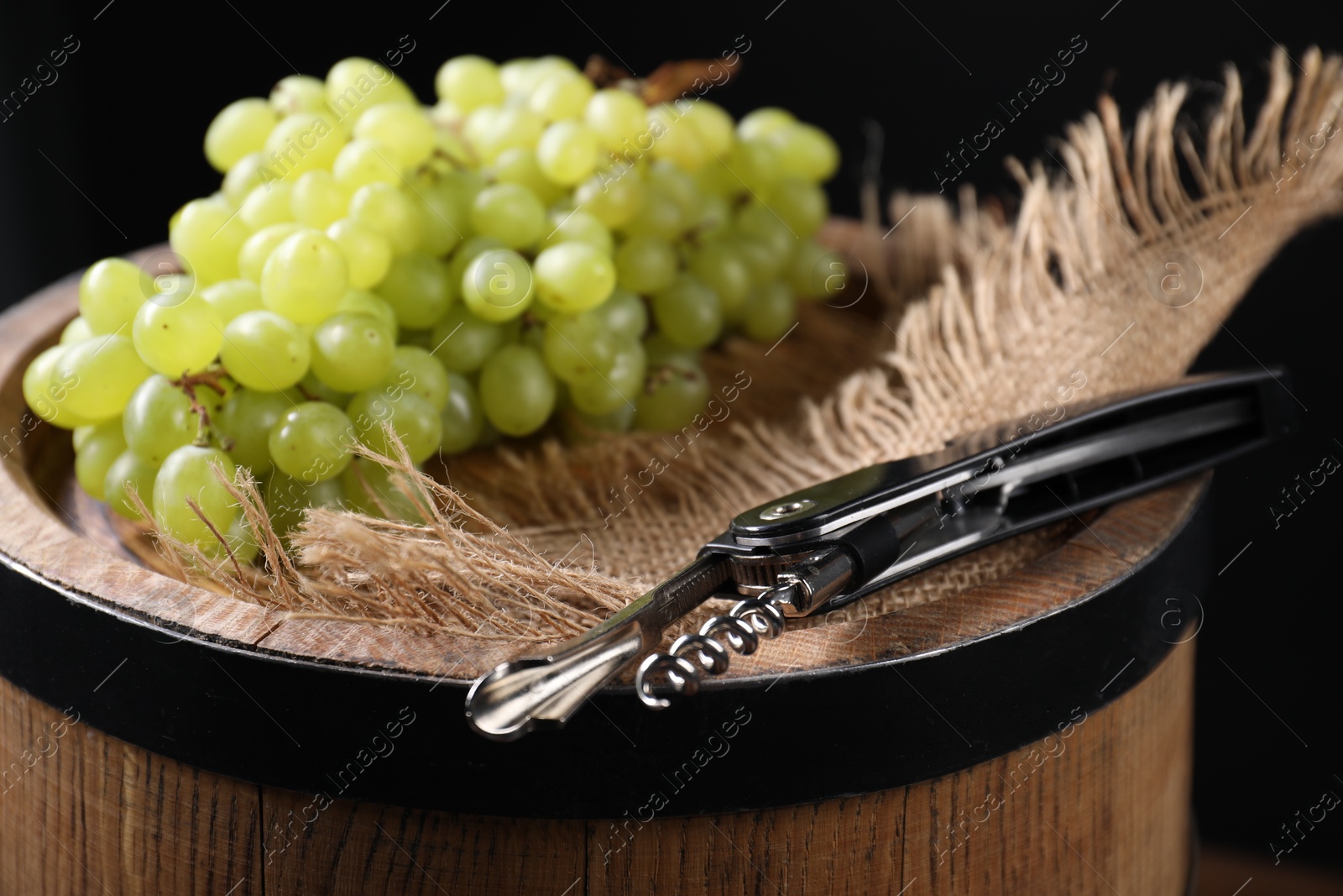 Photo of Sommelier knife with corkscrew and grapes on wooden barrel against black background, closeup