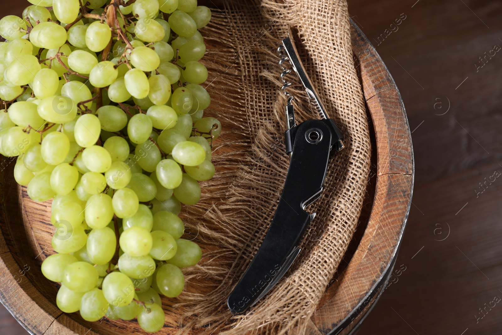 Photo of Sommelier knife with corkscrew, grapes and wooden barrel on table, closeup