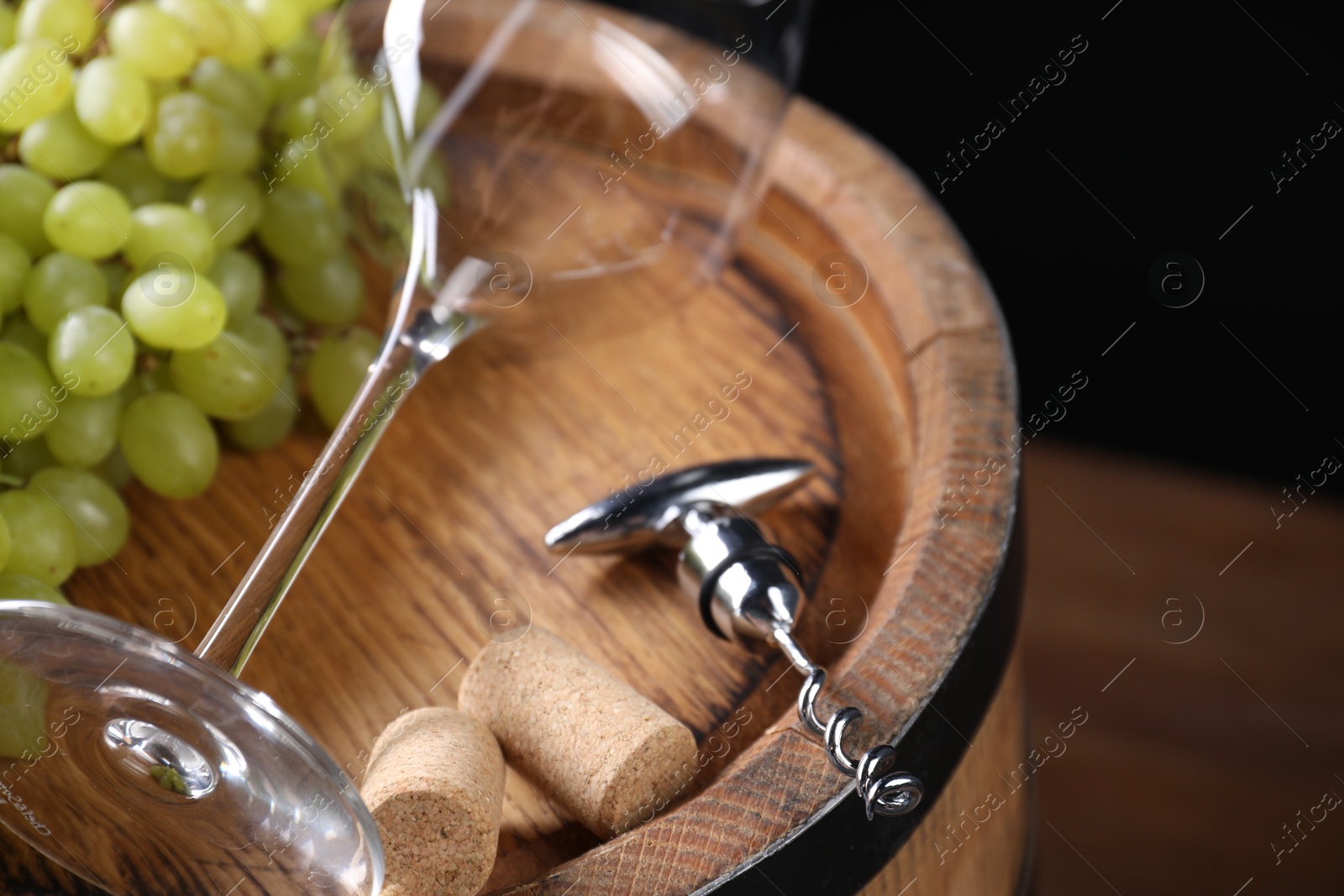 Photo of Corkscrew with metallic handle, glass, corks and grapes on wooden barrel, closeup