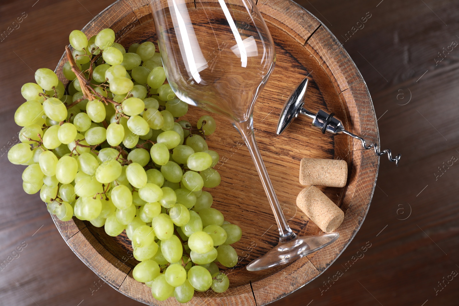 Photo of Corkscrew with metallic handle, glass, corks, grapes and wooden barrel on table, top view