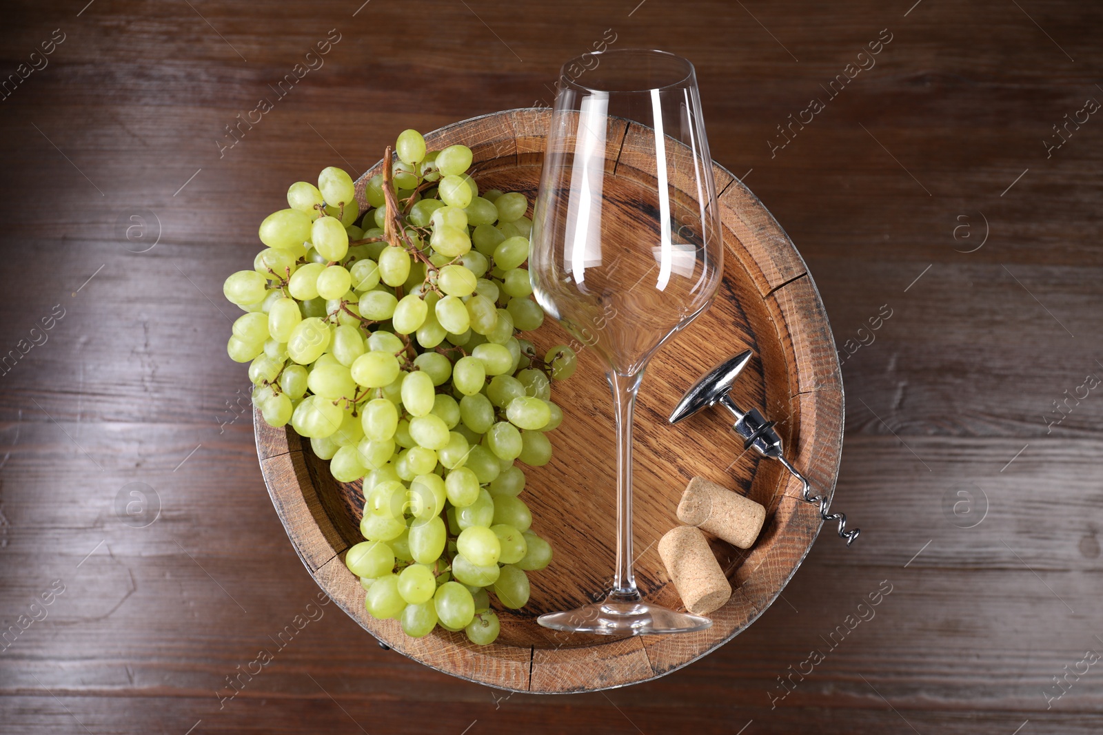 Photo of Corkscrew with metallic handle, glass, corks, grapes and wooden barrel on table, top view