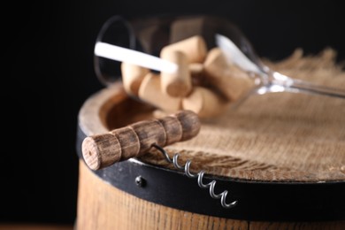 Photo of Corkscrew, corks and glass on wooden barrel against black background, closeup