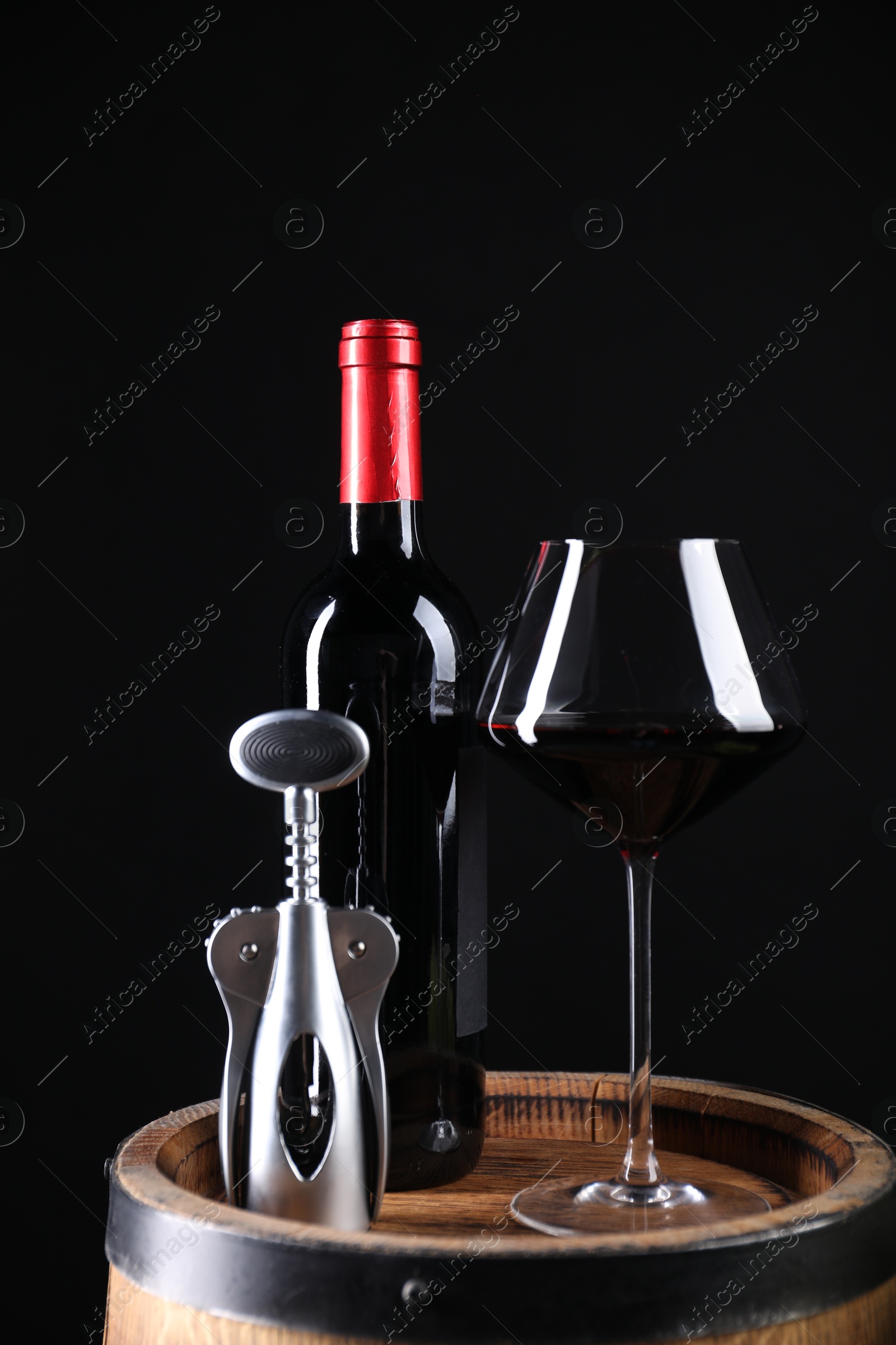 Photo of Wing corkscrew, bottle of wine and glass on wooden barrel against black background