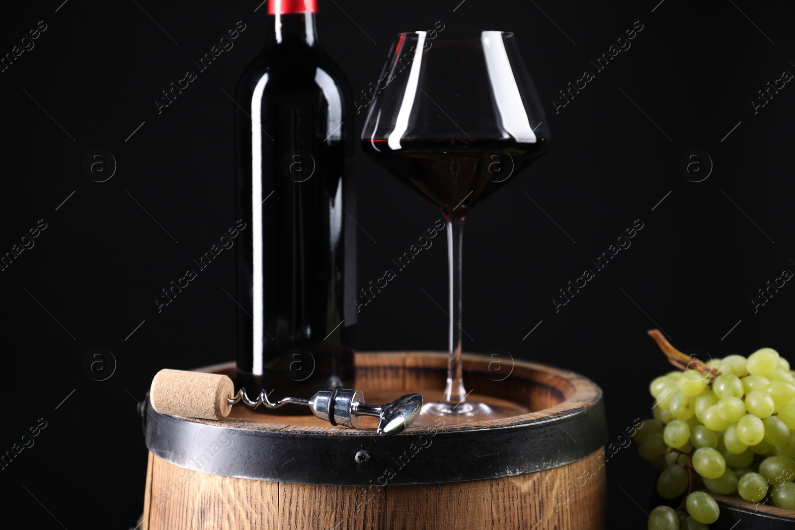 Photo of Corkscrew with metal handle, bottle of wine, glass, grapes and wooden barrel against black background