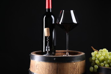 Photo of Corkscrew with metal handle, bottle of wine, glass, grapes and wooden barrel against black background