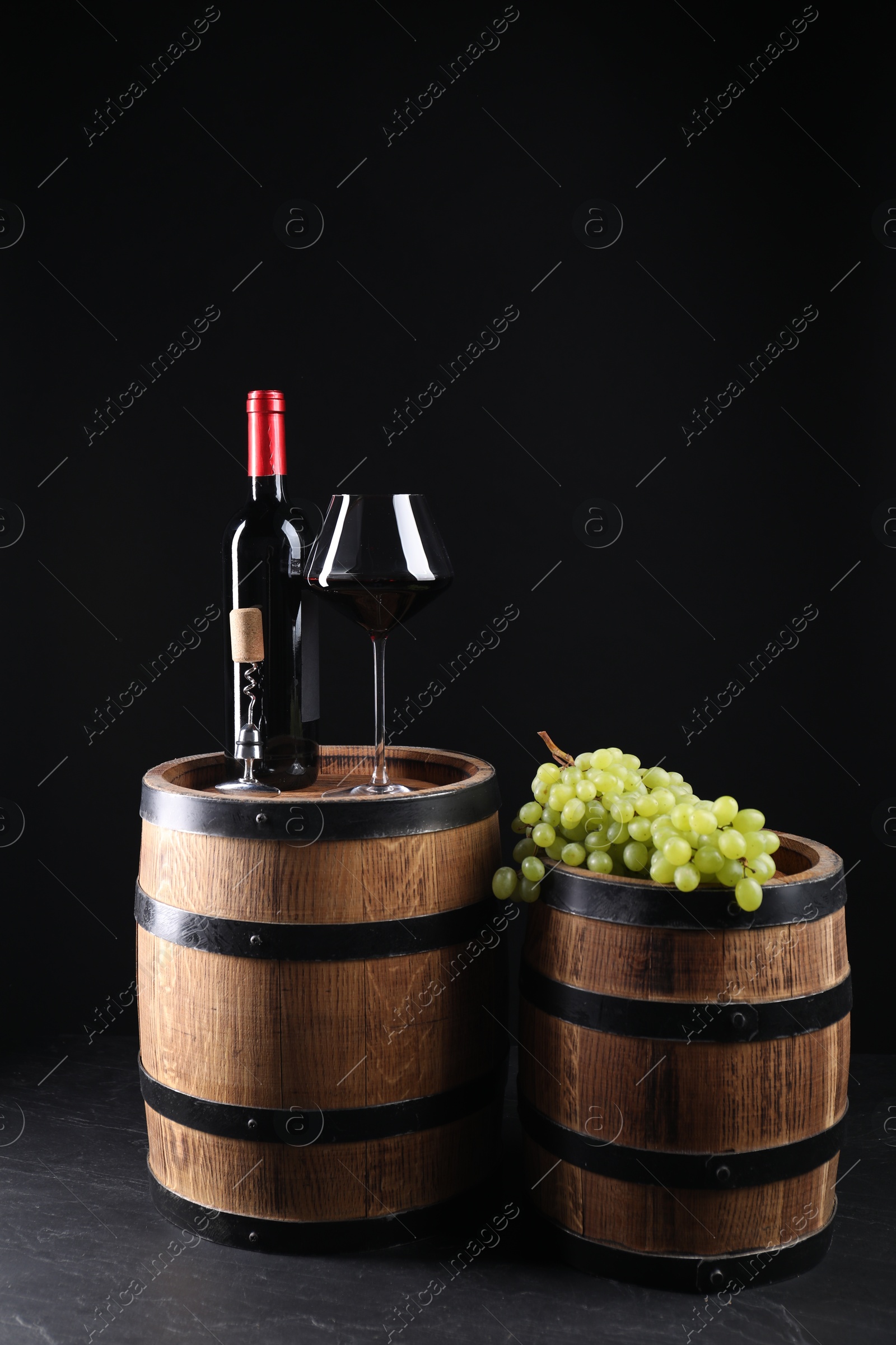 Photo of Corkscrew with metal handle, bottle of wine, glass, grapes and wooden barrels on grey table