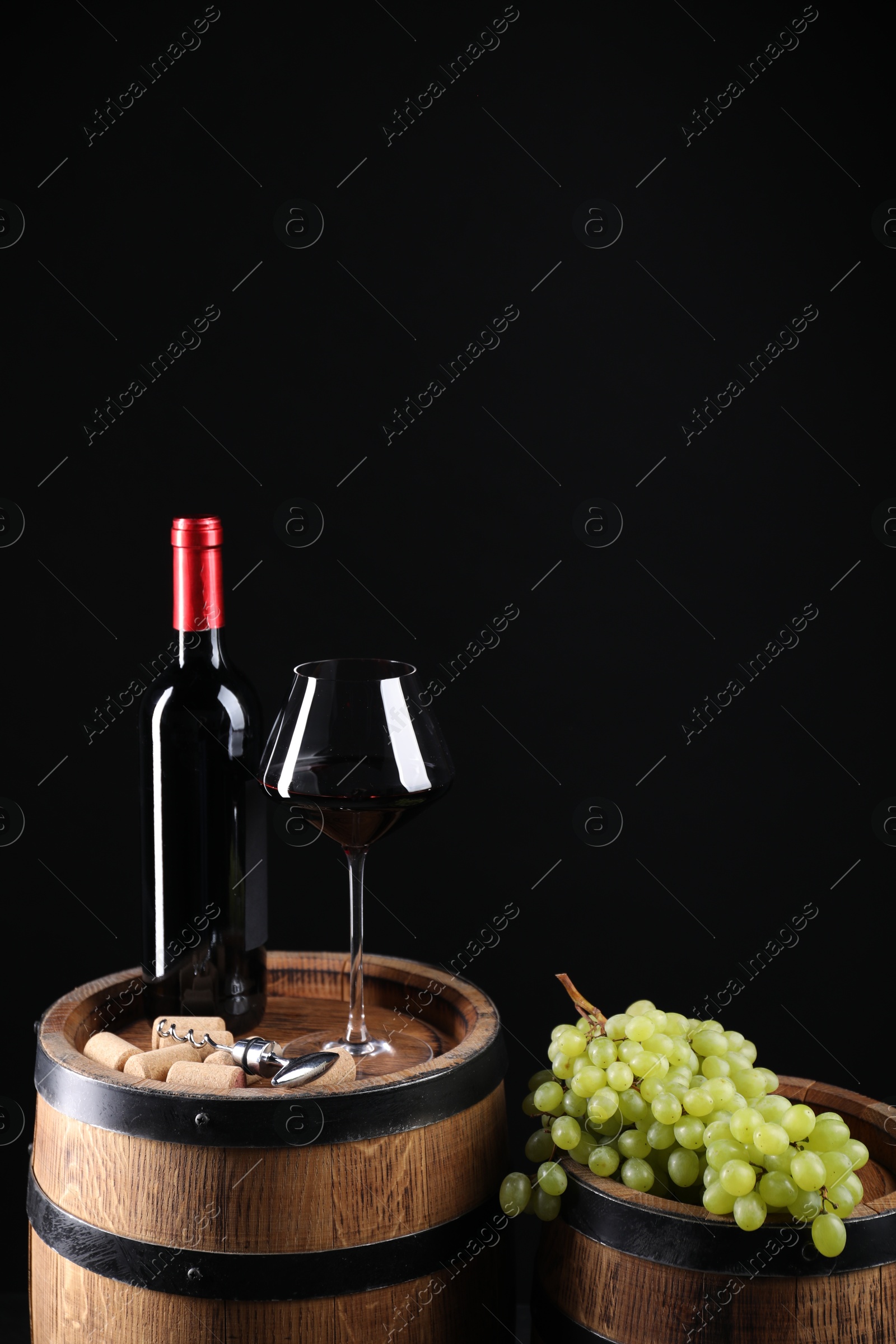 Photo of Corkscrew with metal handle, bottle of wine, glass, grapes and wooden barrels against black background