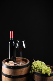 Photo of Corkscrew with metal handle, bottle of wine, glass, grapes and wooden barrels against black background