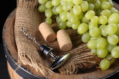 Photo of Corkscrew with metal handle, corks and grapes on wooden barrel, closeup