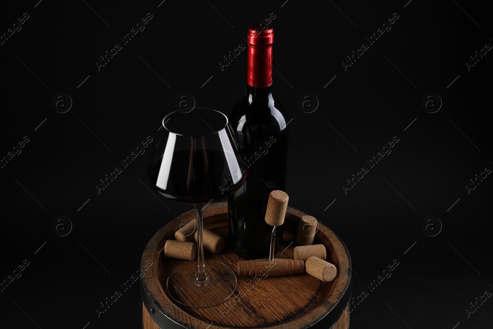 Photo of Corkscrew, bottle of wine, glass and corks on wooden barrel against black background