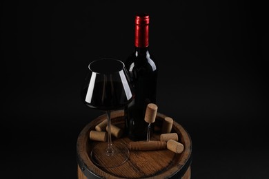 Photo of Corkscrew, bottle of wine, glass and corks on wooden barrel against black background