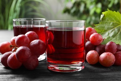 Photo of Tasty grape juice in glasses and berries on black wooden table against blurred green background, closeup