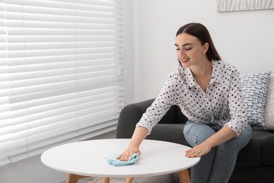 Photo of Beautiful young woman wiping coffee table with rag at home