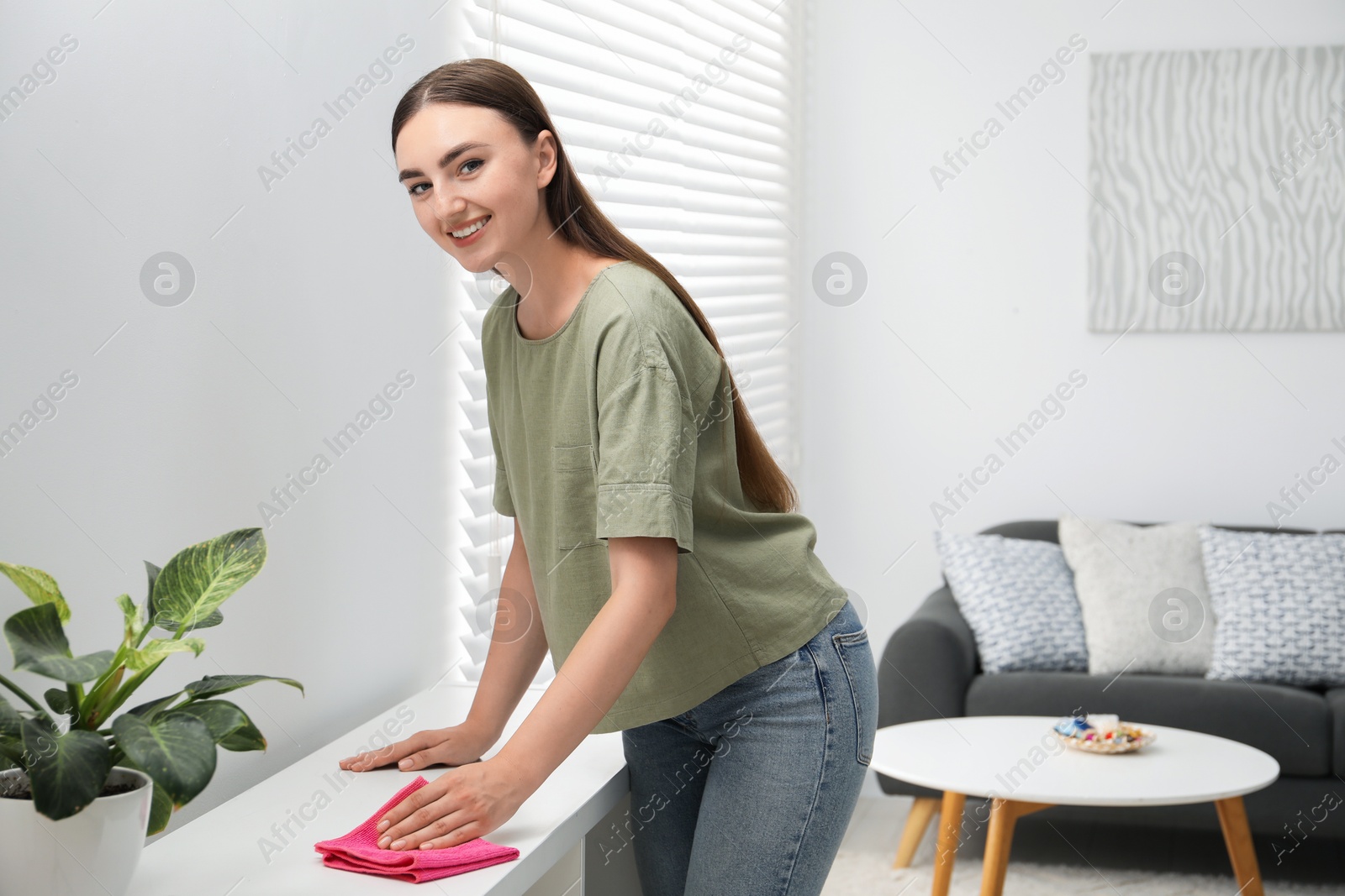 Photo of Beautiful young woman wiping furniture with rag at home