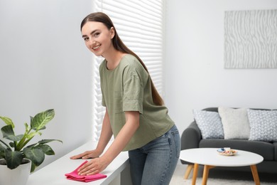 Photo of Beautiful young woman wiping furniture with rag at home