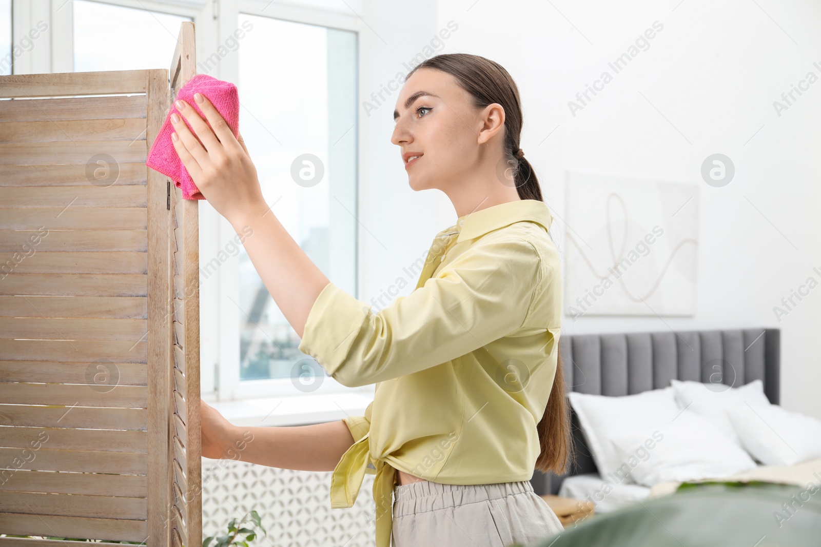 Photo of Beautiful young woman wiping folding screen with rag in bedroom